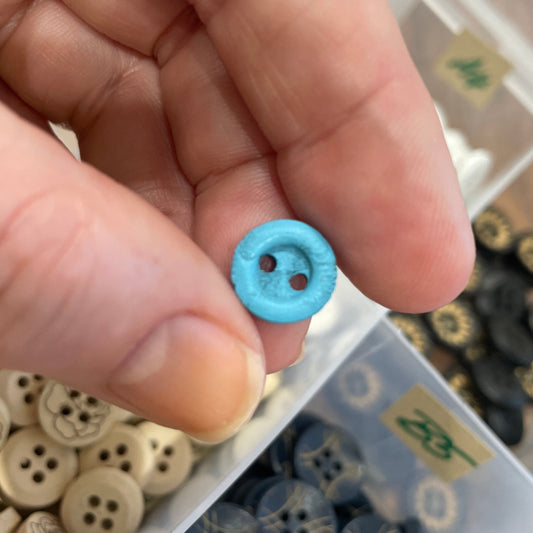 Hand holding a small blue button with a container of various buttons in the background.