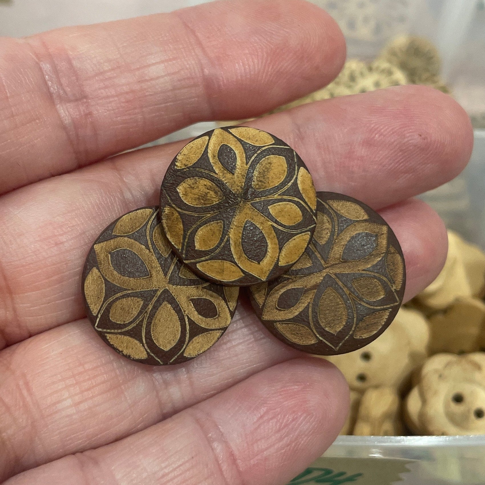 Hand holding three wooden buttons with floral patterns in front of a container filled with various buttons.