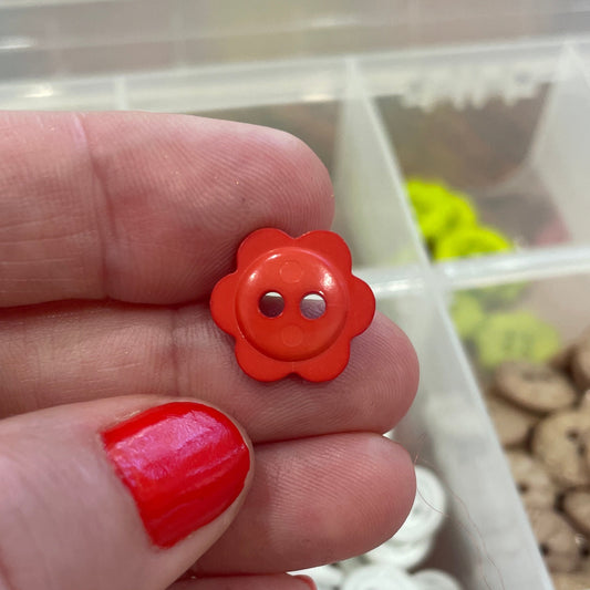 Hand holding a red flower-shaped button with a background of button collection.