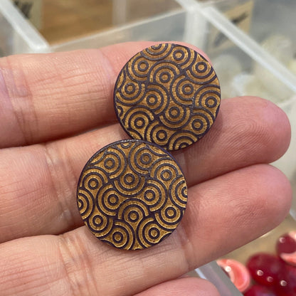 Two wooden buttons with a geometric pattern held between fingers, with a clear container in the background.
