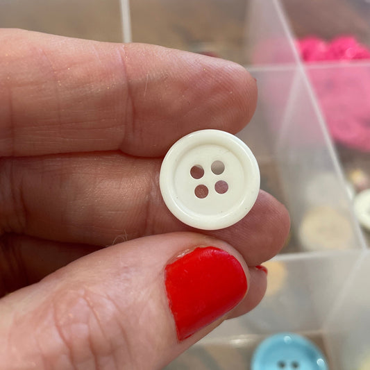 Hand holding a white button with four holes in front of a container of colorful buttons.