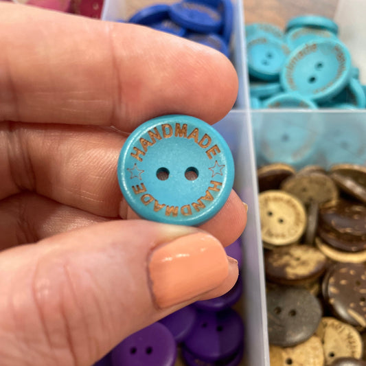 Hand holding a blue button with 'Handmade' text, surrounded by various buttons in a box.