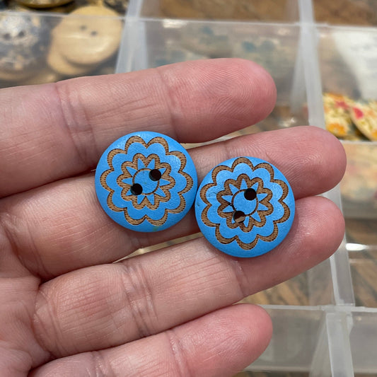 Hand holding two blue buttons with floral patterns in front of a container of assorted buttons.