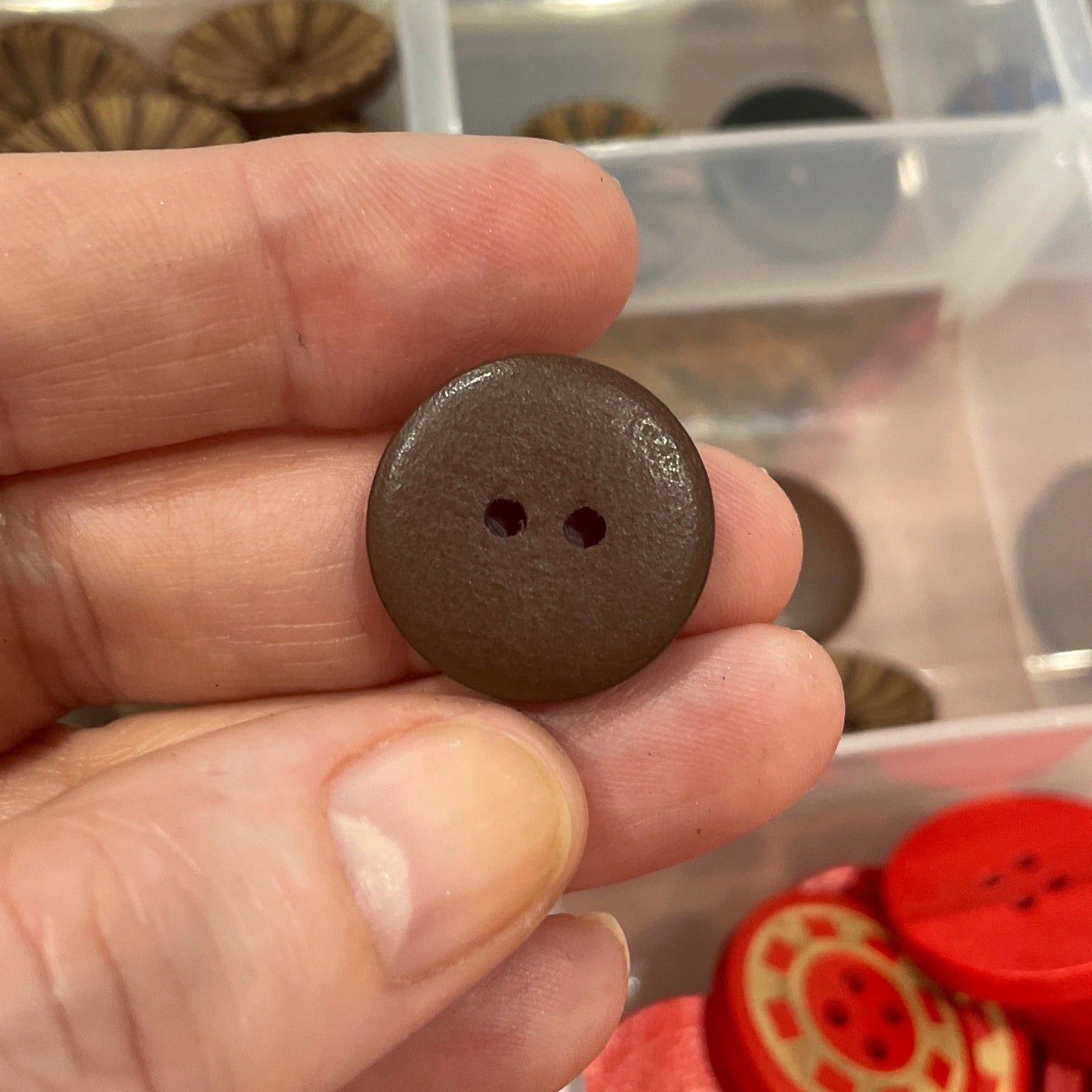 Hand holding a brown button with a clear container of assorted buttons in the background