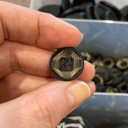 Hand holding a small decorative button with intricate design, surrounded by other buttons in a container.
