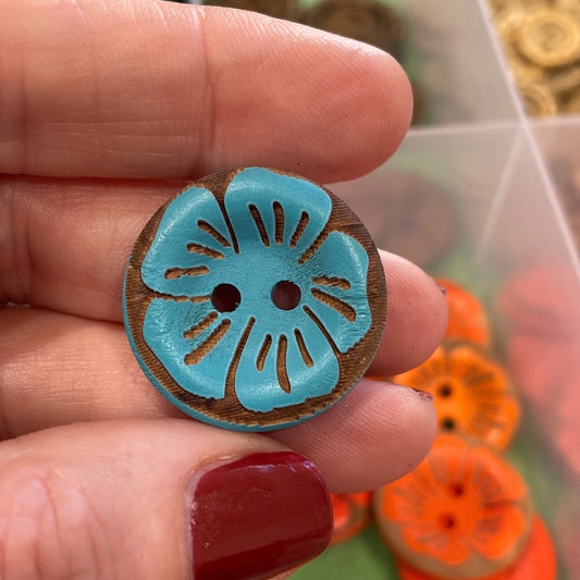 Hand holding a decorative button with a blue flower design, against a background of colorful buttons.