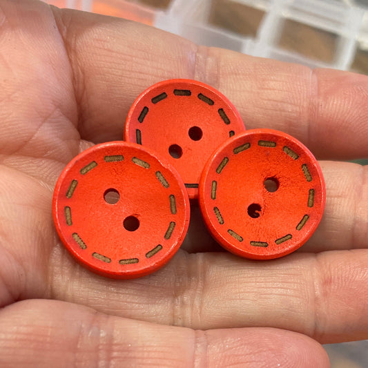 Hand holding three orange buttons with a container of assorted buttons in the background.