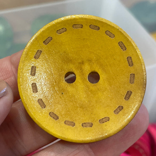 Hand holding a yellow button with a patterned edge, against a background of various buttons in a container.