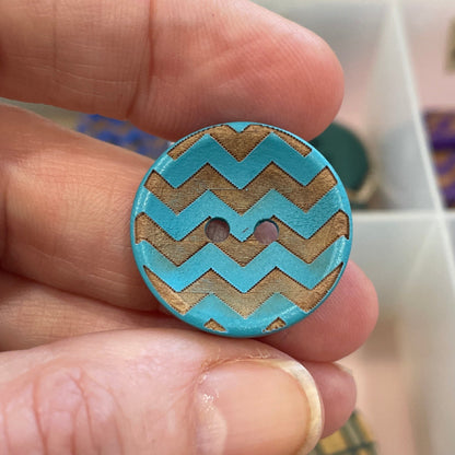 Hand holding a blue and gold zigzag patterned button with a container of buttons in the background.