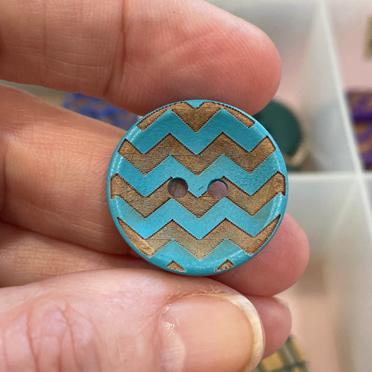 Hand holding a blue and gold zigzag patterned button with a container of buttons in the background.