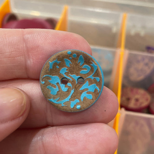 Hand holding a decorative button with blue patterns against a blurred background of a button collection.