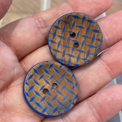 Two wooden buttons with blue pattern held in a hand, with a container of similar buttons in the background.