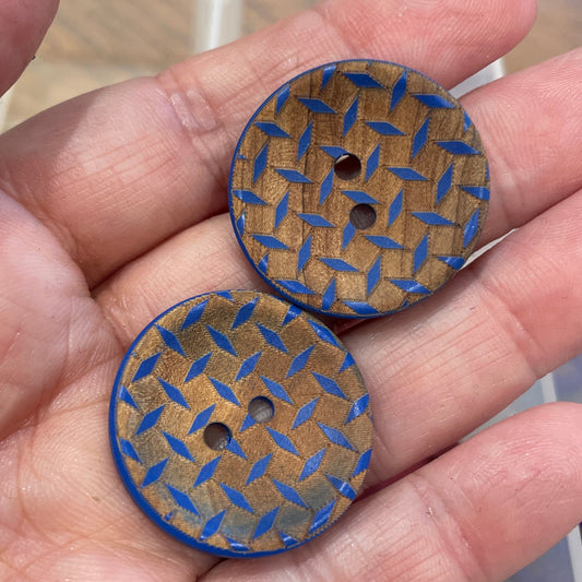 Two wooden buttons with blue pattern held in a hand, with a container of similar buttons in the background.