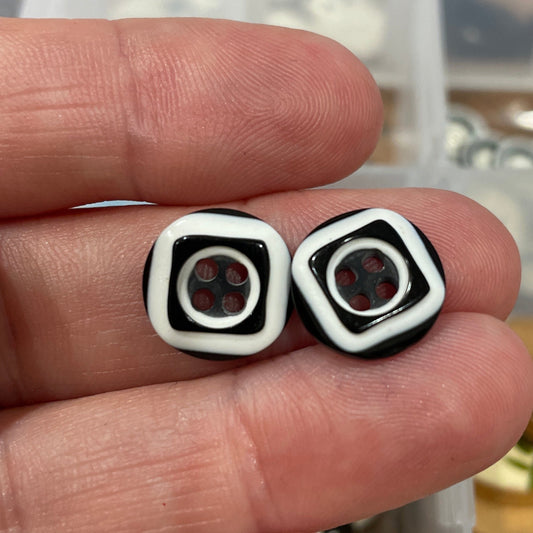 Black and white square-shaped earrings held between fingers with a container of buttons in the background.