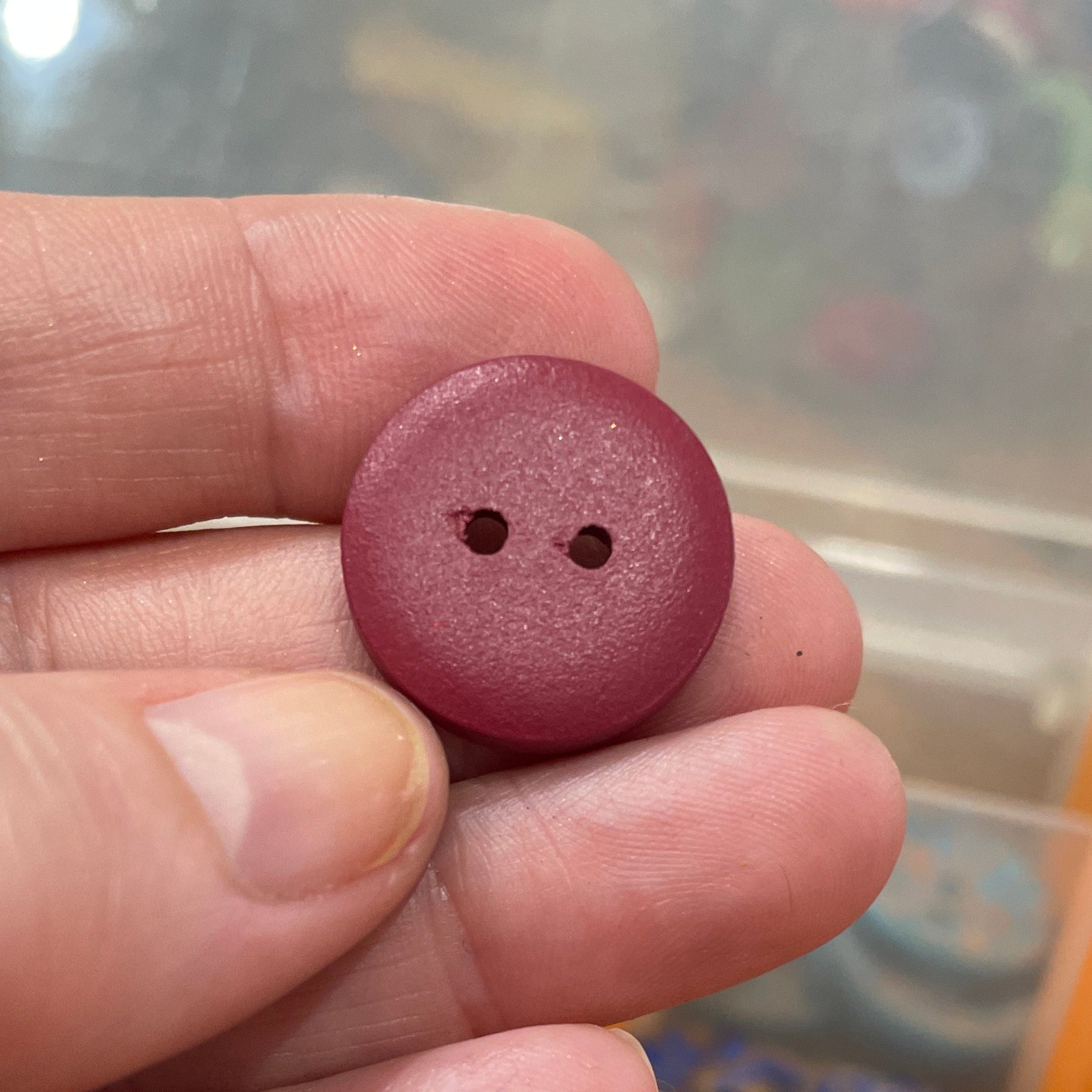 Maroon button held between fingers with a container of buttons in the background
