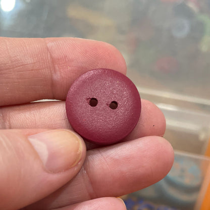 Maroon button held between fingers with a container of buttons in the background