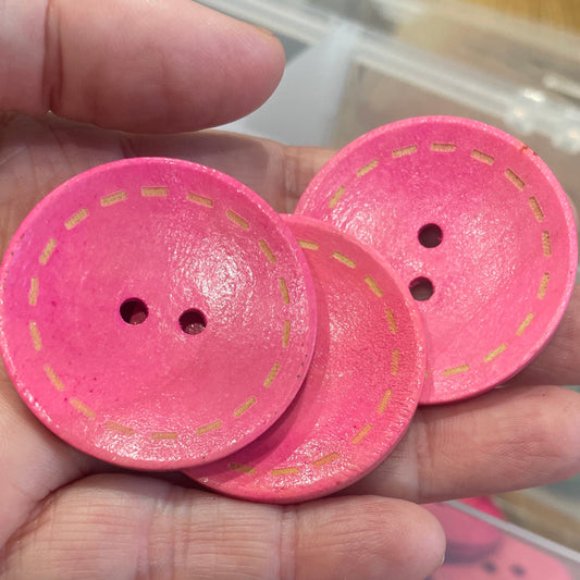 Pink buttons with a hand holding two buttons above a container filled with pink buttons.
