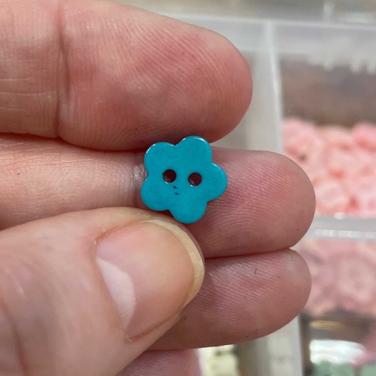 Hand holding a blue flower-shaped button with a background of button storage containers.