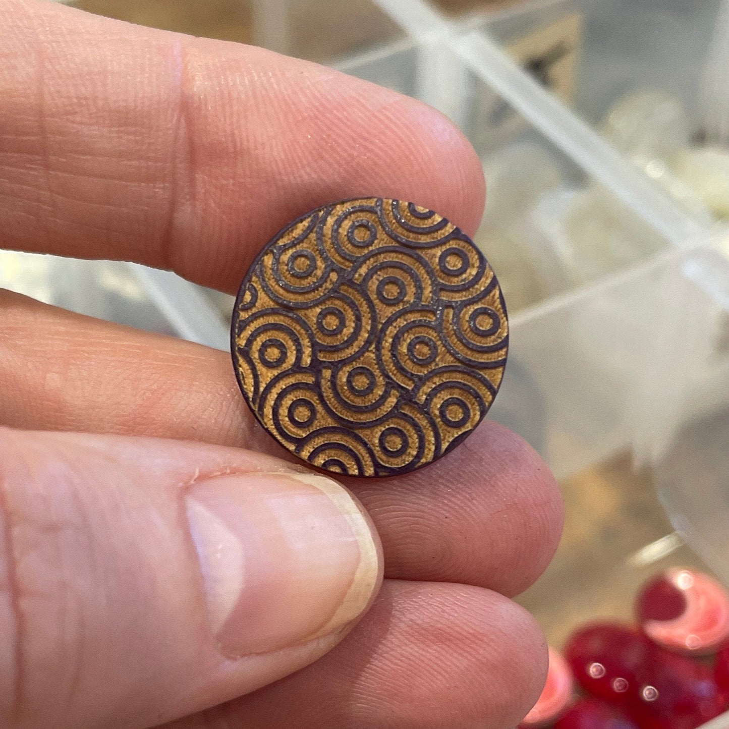 Hand holding a decorative button with a detailed pattern, blurred background of a storage box with various items.