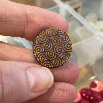 Hand holding a decorative button with a detailed pattern, blurred background of a storage box with various items.