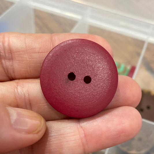 Hand holding a maroon button with a clear container of similar buttons in the background