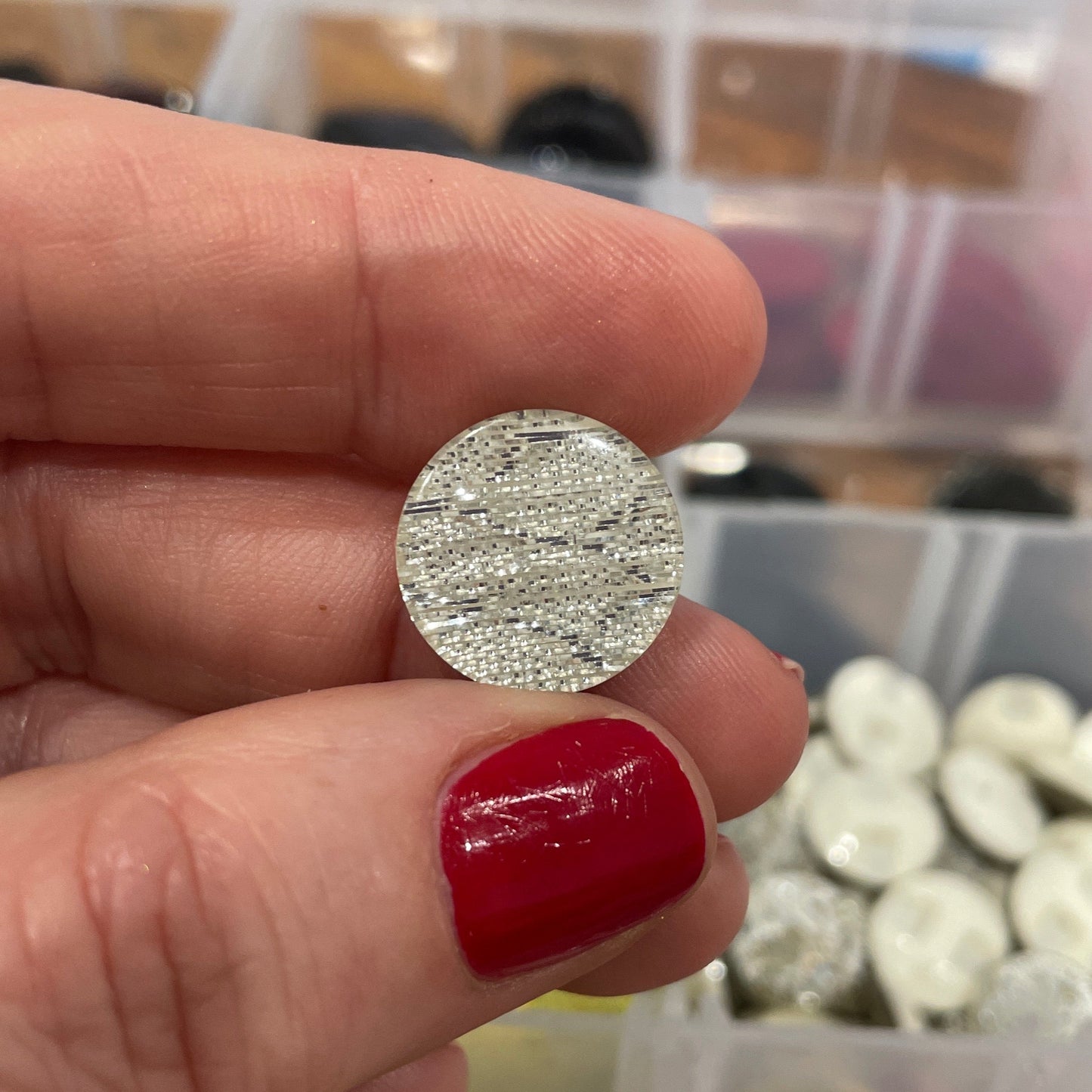 Silver round earring held between fingers with a blurred background of colorful buttons.