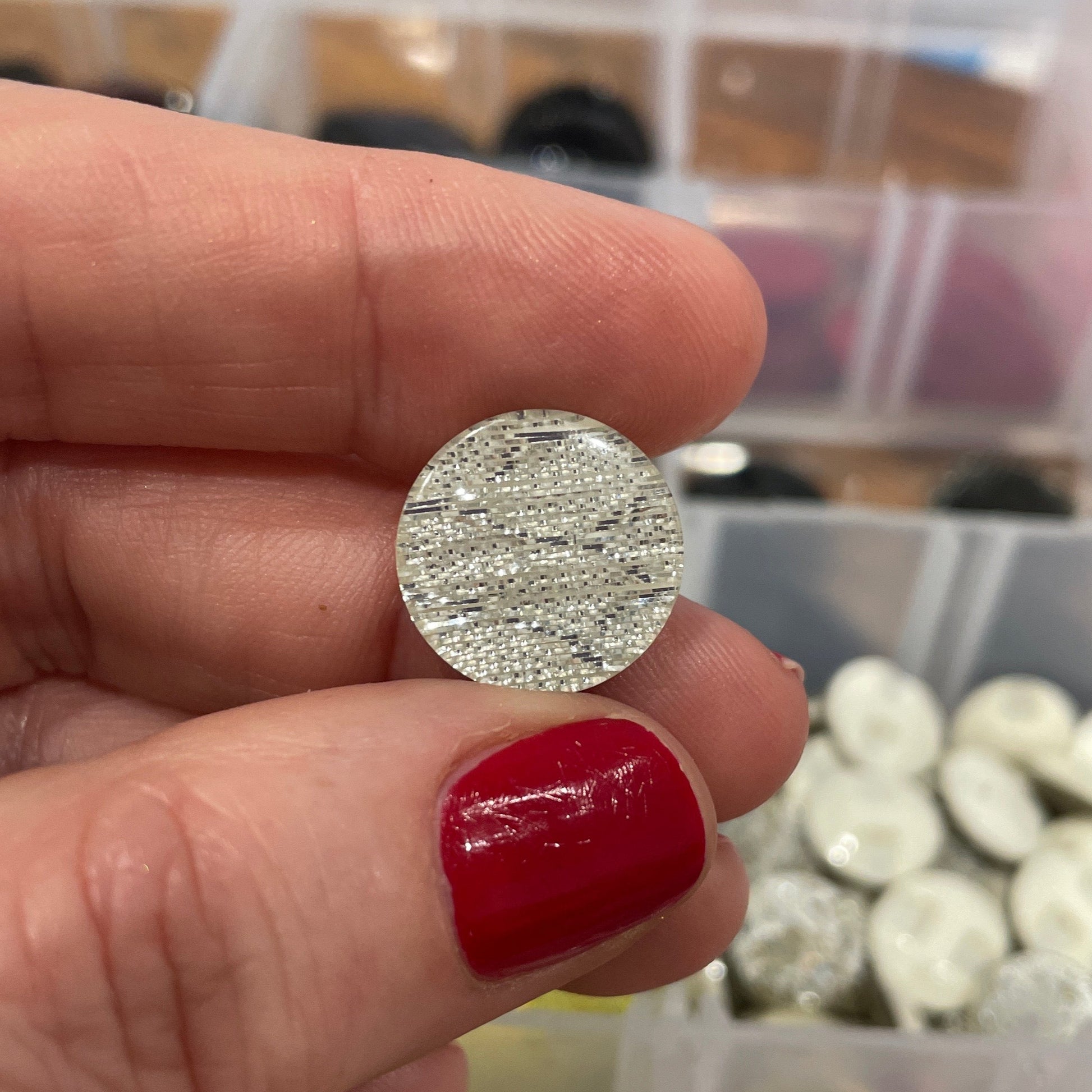 Silver round earring held between fingers with a blurred background of colorful buttons.