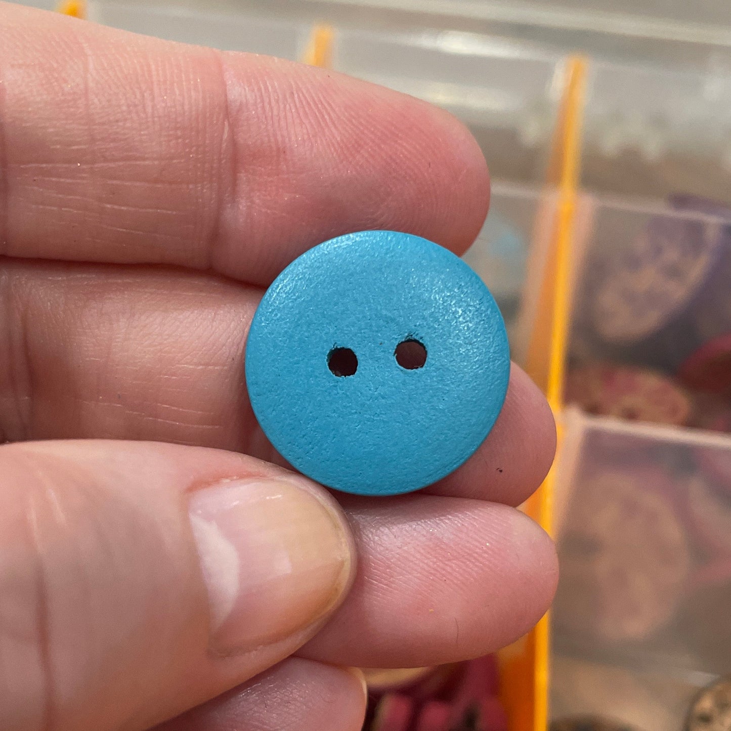 Hand holding a blue button with a blurred background of a button collection box.