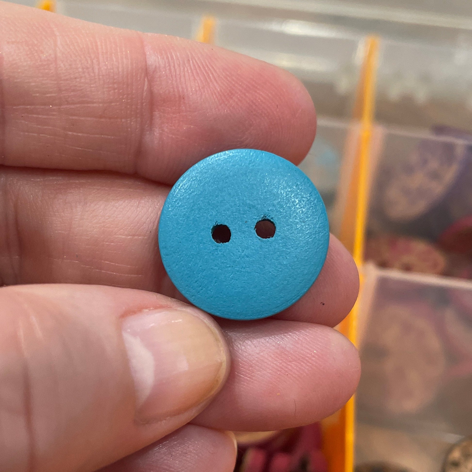 Hand holding a blue button with a blurred background of a button collection box.