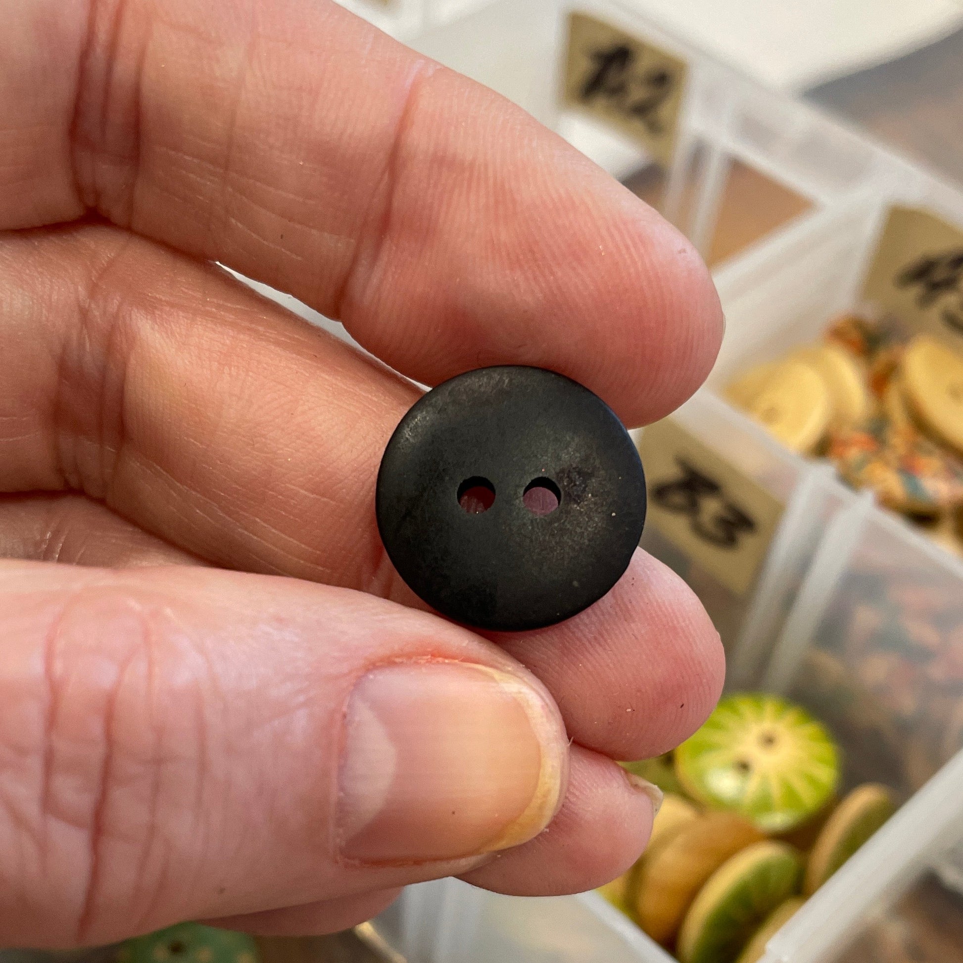 Hand holding a black button with a container of assorted buttons in the background