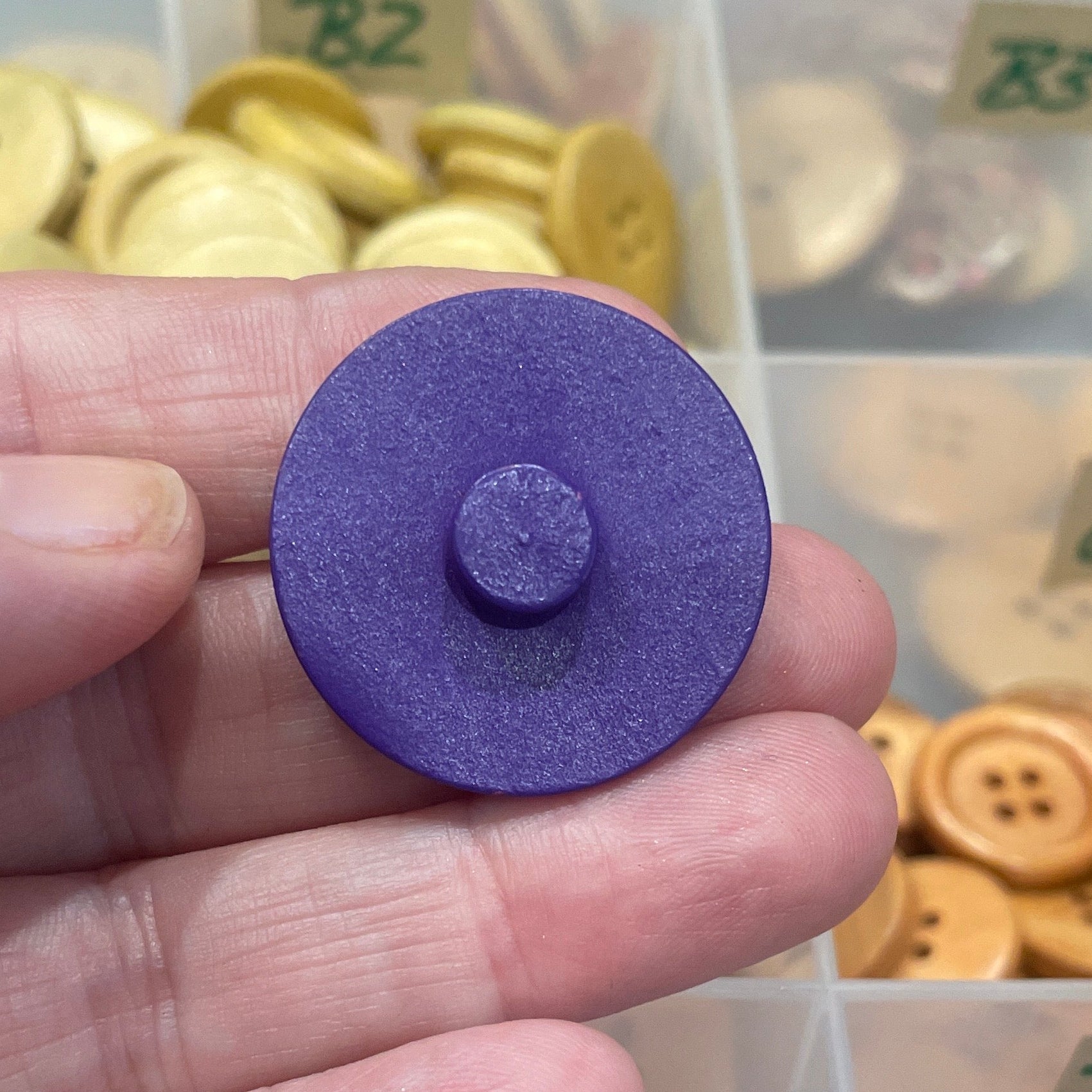 Hand holding a purple button with a container of buttons in the background