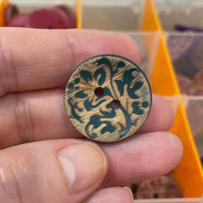 Hand holding a decorative button with intricate design in front of an open container with buttons.