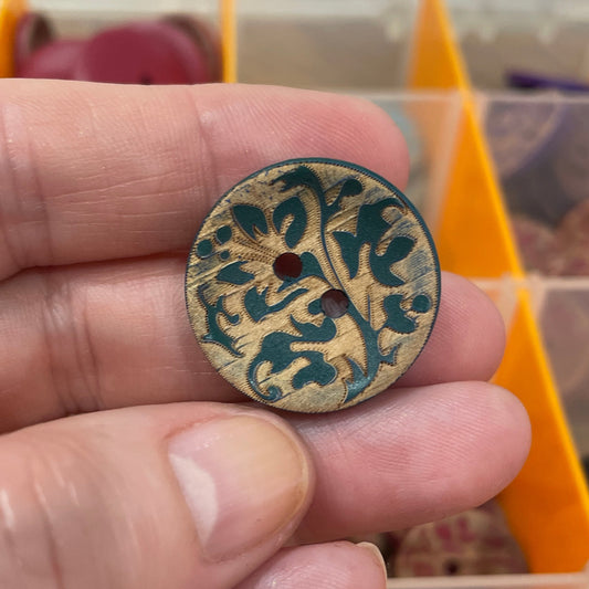 Hand holding a decorative button with intricate design in front of an open container with buttons.