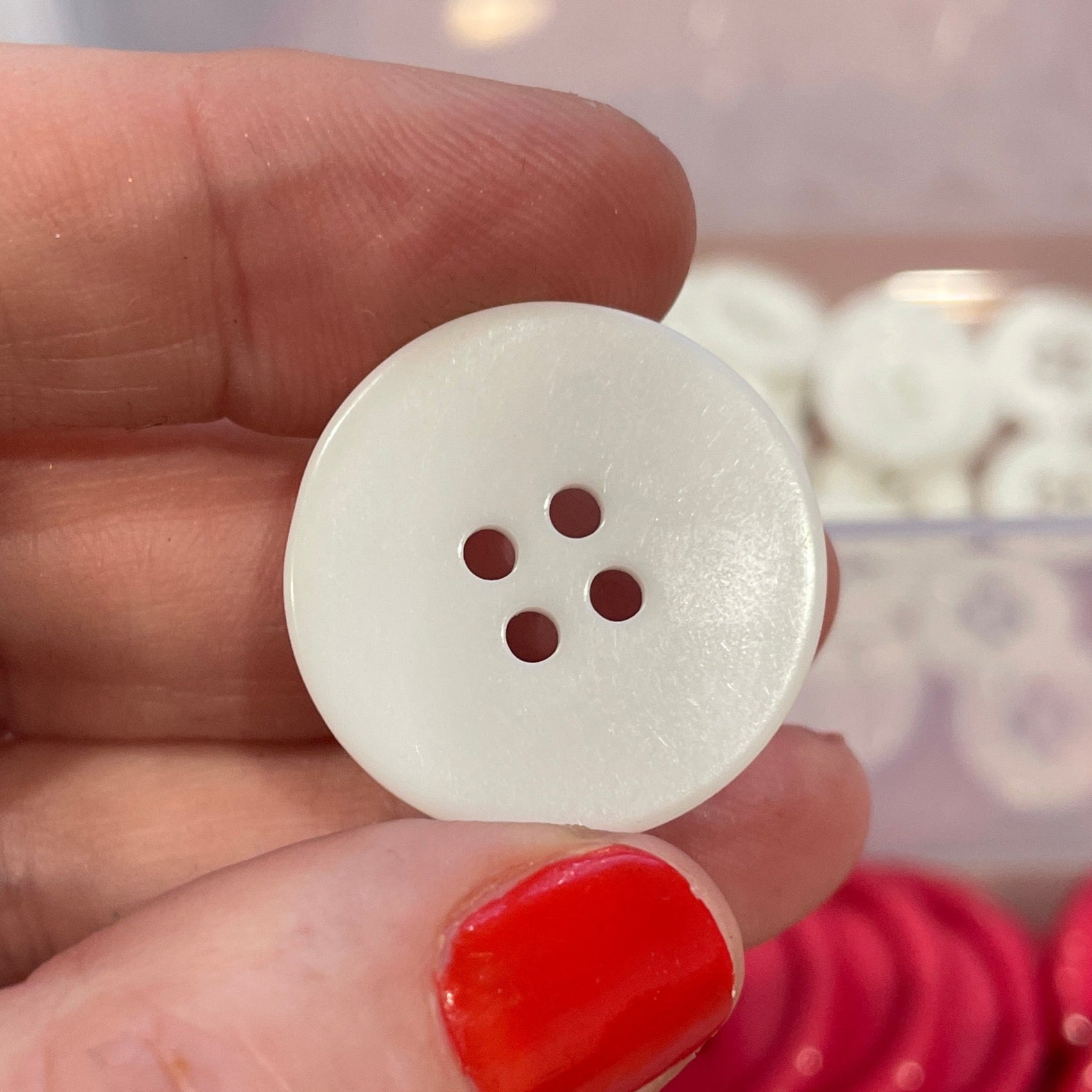 White button held by a hand with red nail polish, against a background of red buttons in a container.