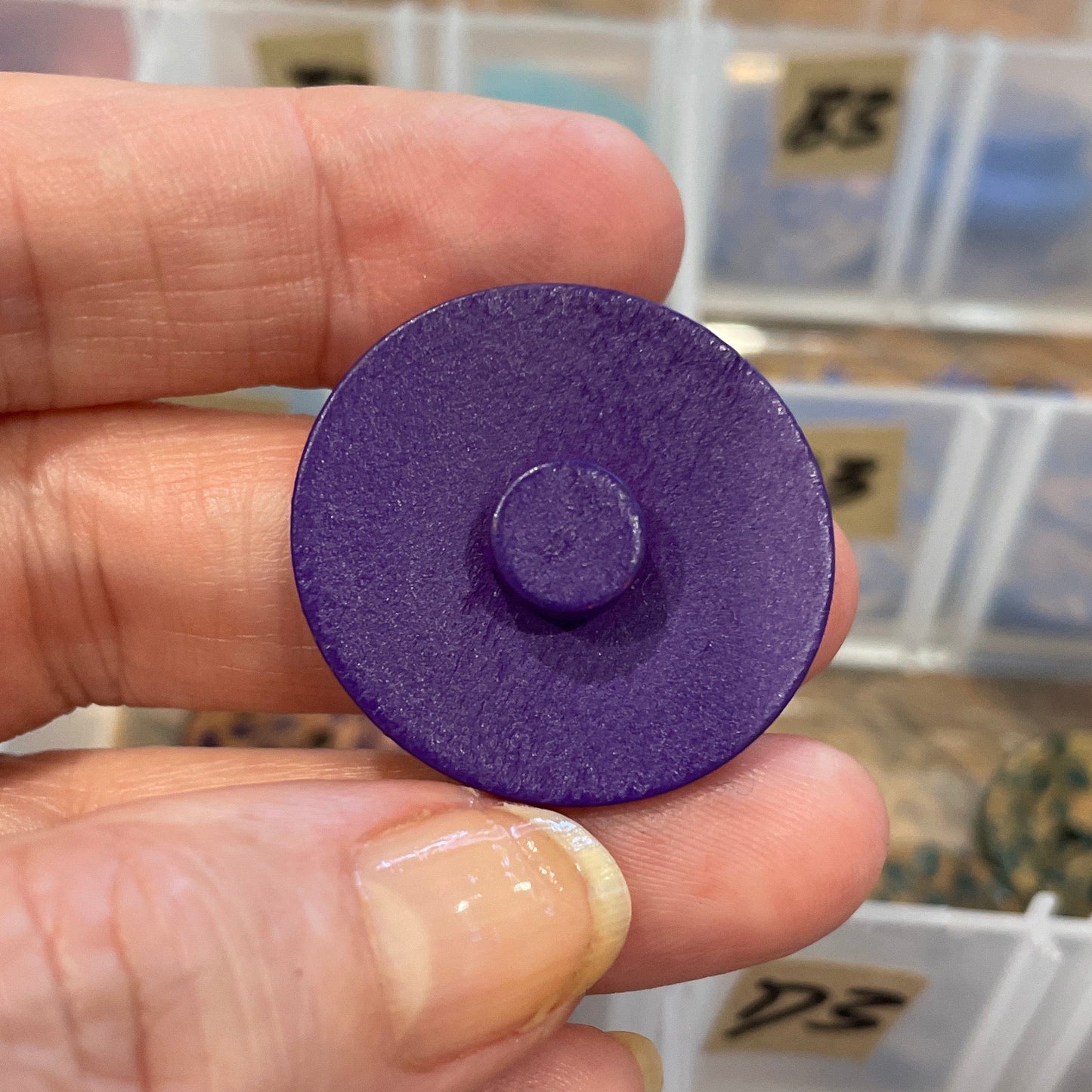 Hand holding a purple button with a background of button storage containers.