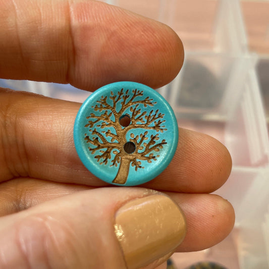Hand holding a blue button with a tree design against a background of a clear container with buttons.