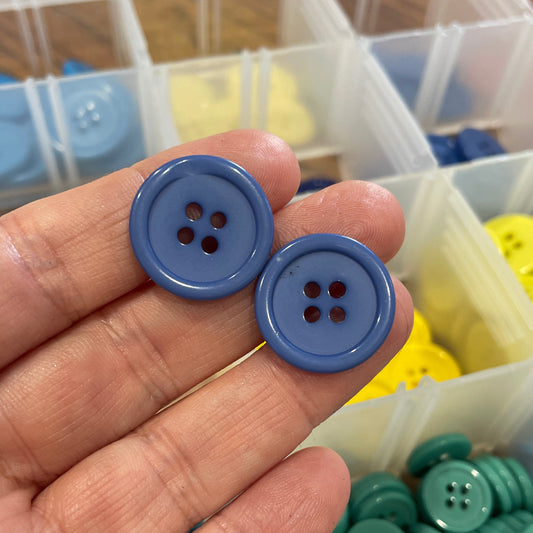 Hand holding two blue buttons with a container of colorful buttons in the background