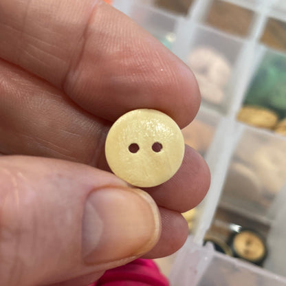 Hand holding a yellow button with a blurred background of assorted buttons in a container.
