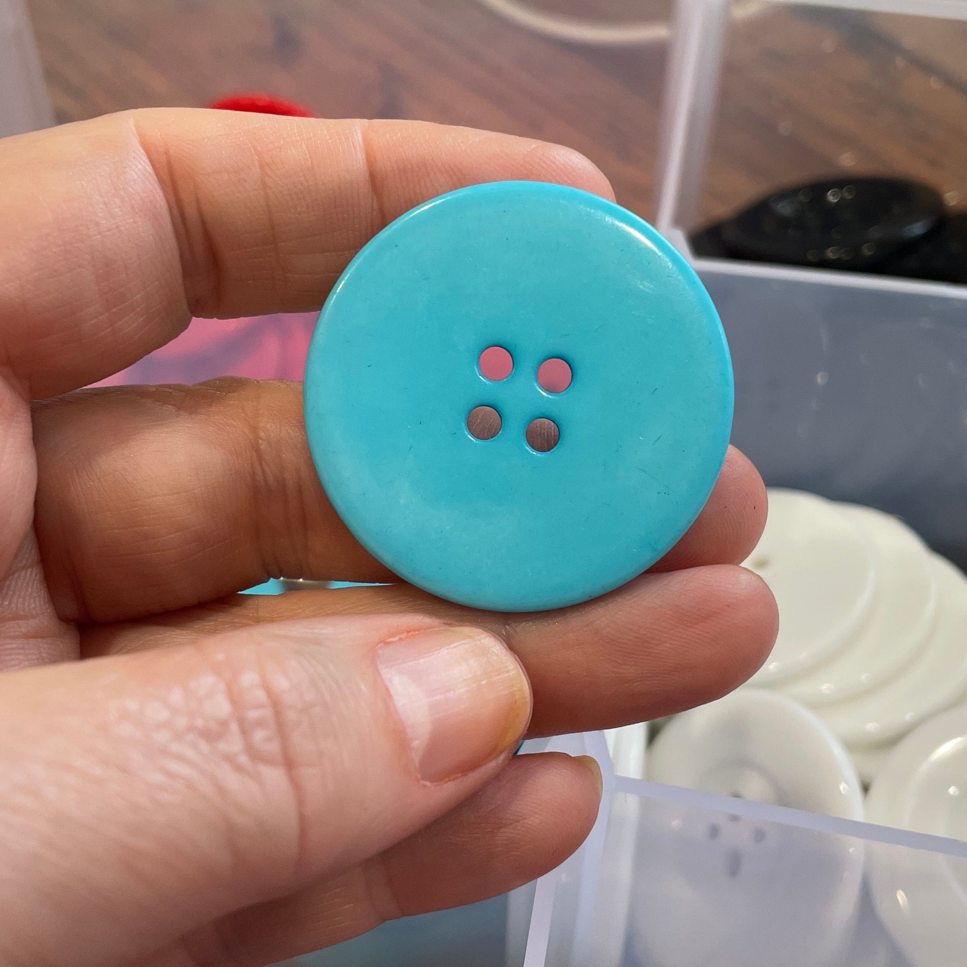 Hand holding a blue button with four holes against a blurred background of buttons and containers.