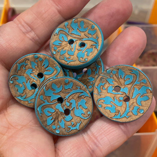 Hand holding decorative buttons with intricate designs, with a box of buttons in the background.