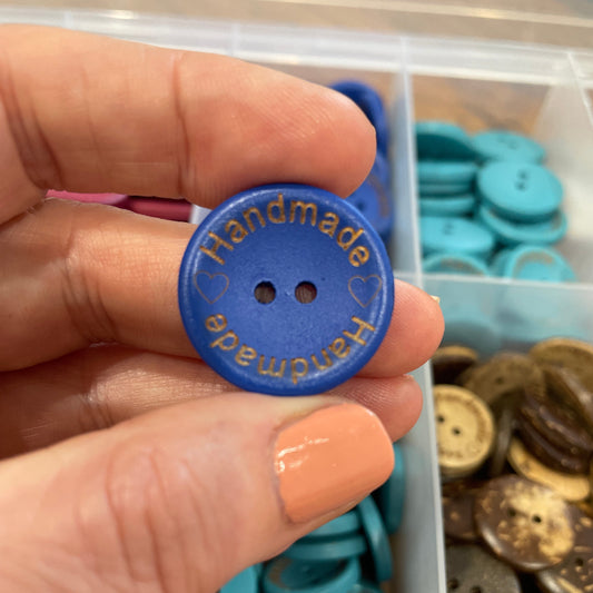 Hand holding a blue button with 'Handmade' text, against a background of assorted buttons.