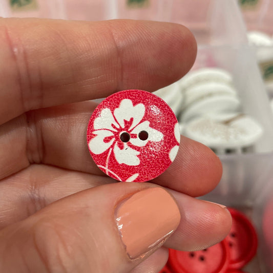 Hand holding a red button with white floral design, with a container of buttons in the background.