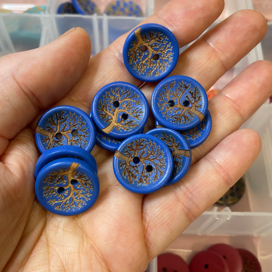 Hand holding blue buttons with tree design, with a container of buttons in the background.