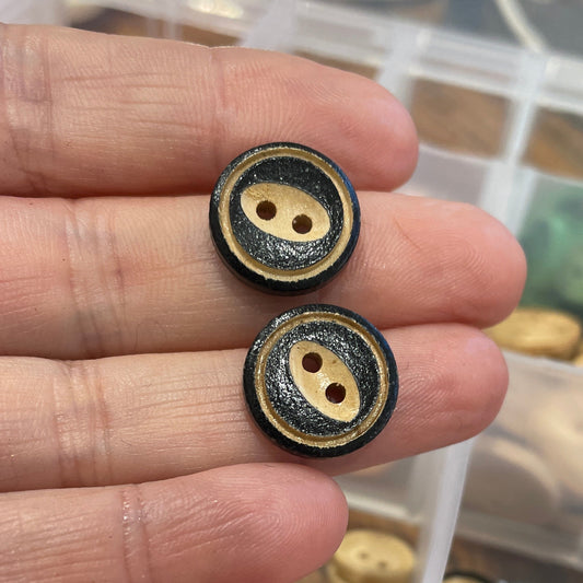 Two black and gold buttons held between fingers with a container of assorted buttons in the background.