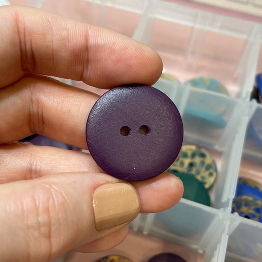 Hand holding a purple button with a container of buttons in the background