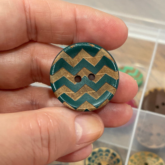 Hand holding a decorative button with a zigzag pattern over a container of assorted buttons.