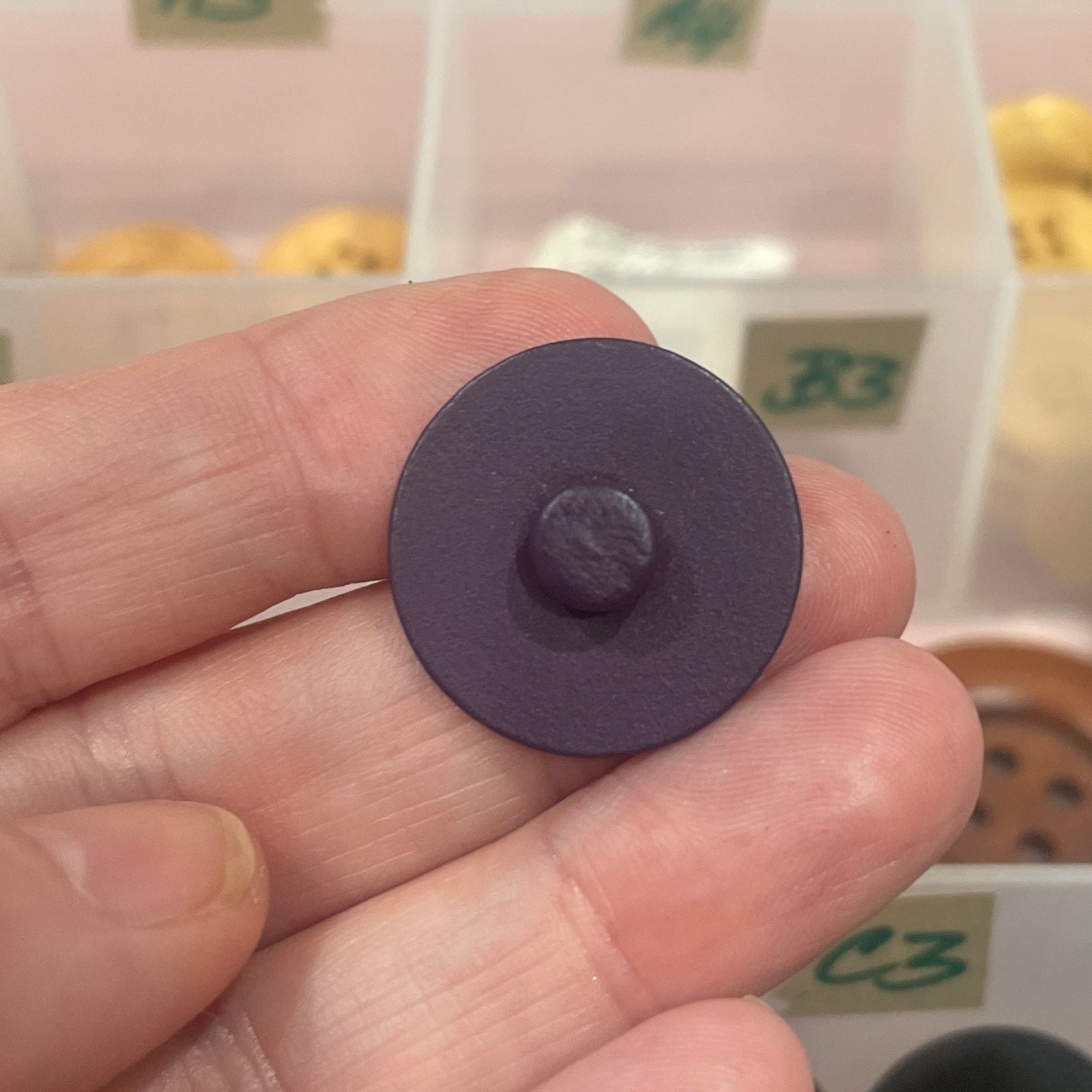 Hand holding a purple button with a container of buttons in the background