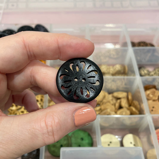Hand holding a black decorative button with a detailed center, against a background of a clear container with various buttons.