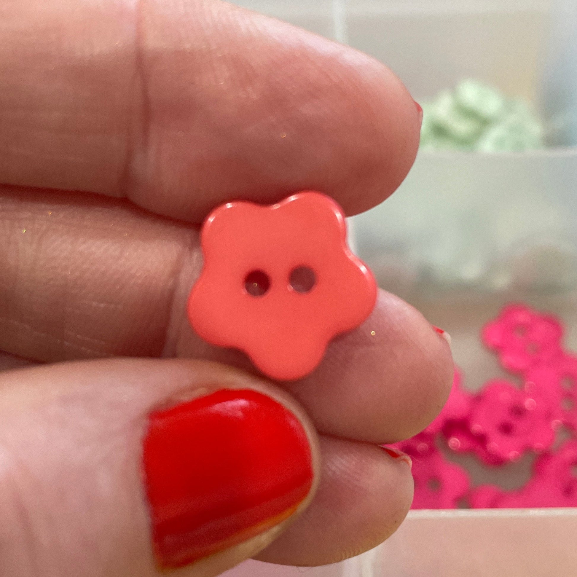 Hand holding a red flower-shaped button with a blurred background of colorful buttons in a box.