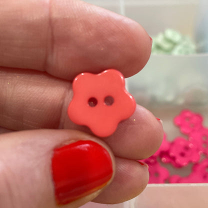 Hand holding a red flower-shaped button with a blurred background of colorful buttons in a box.